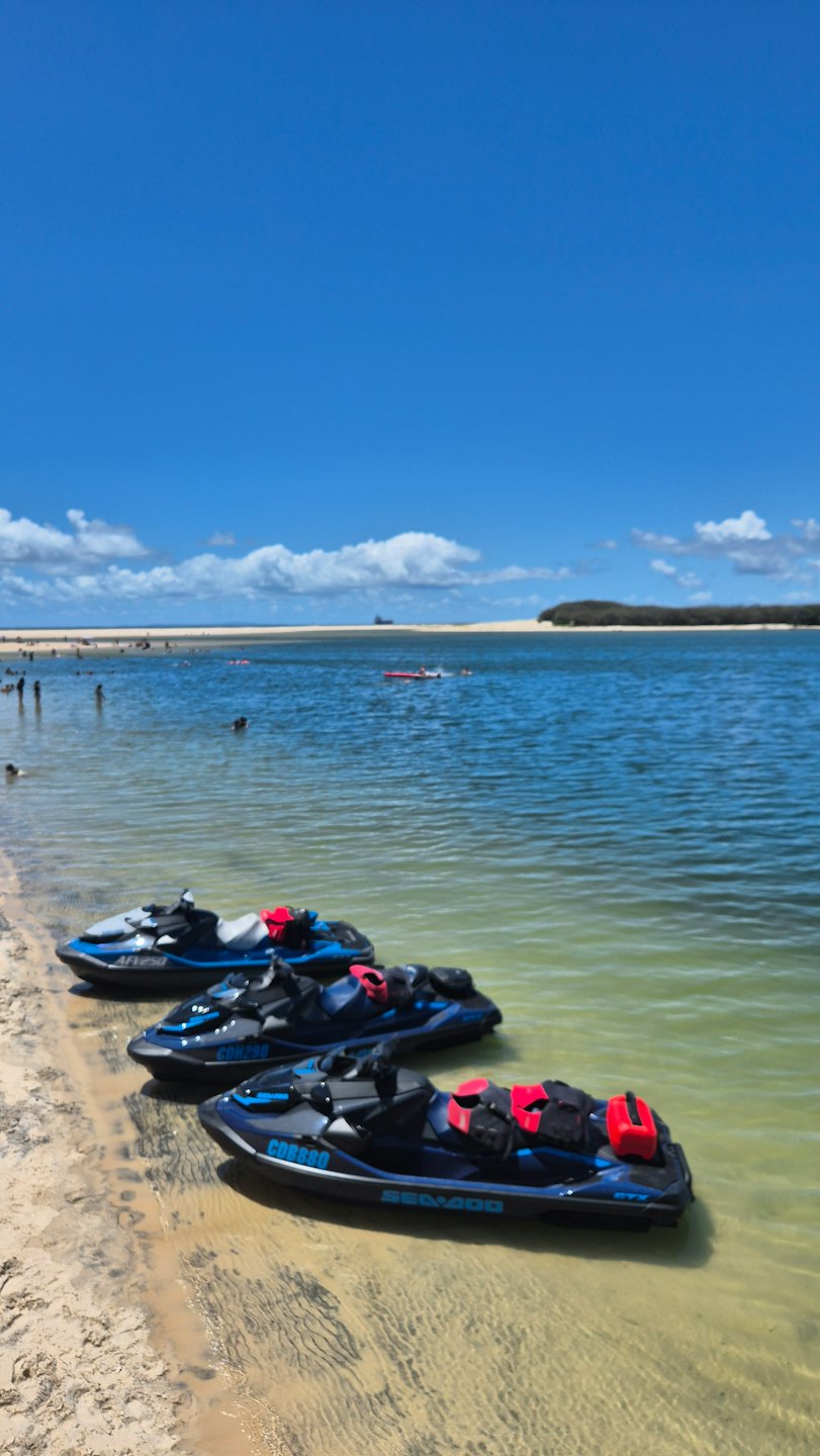 Three jet skis rest on a sandy beach near water.
